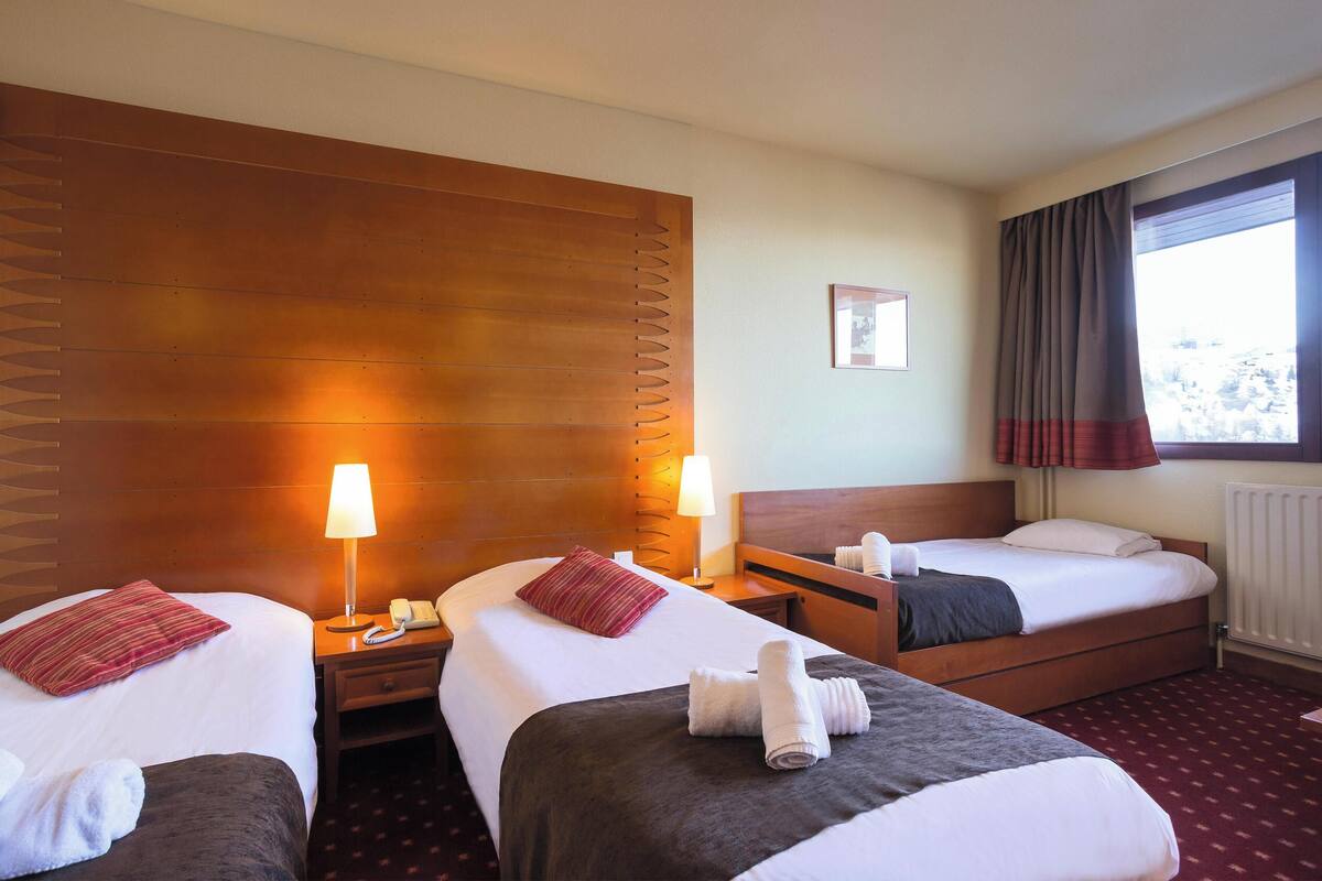 Ein Hotelzimmer mit zwei Betten, einem hölzernen Kopfteil, einem Nachttisch mit Lampe, einem Fenster mit Vorhängen und einer Heizung.