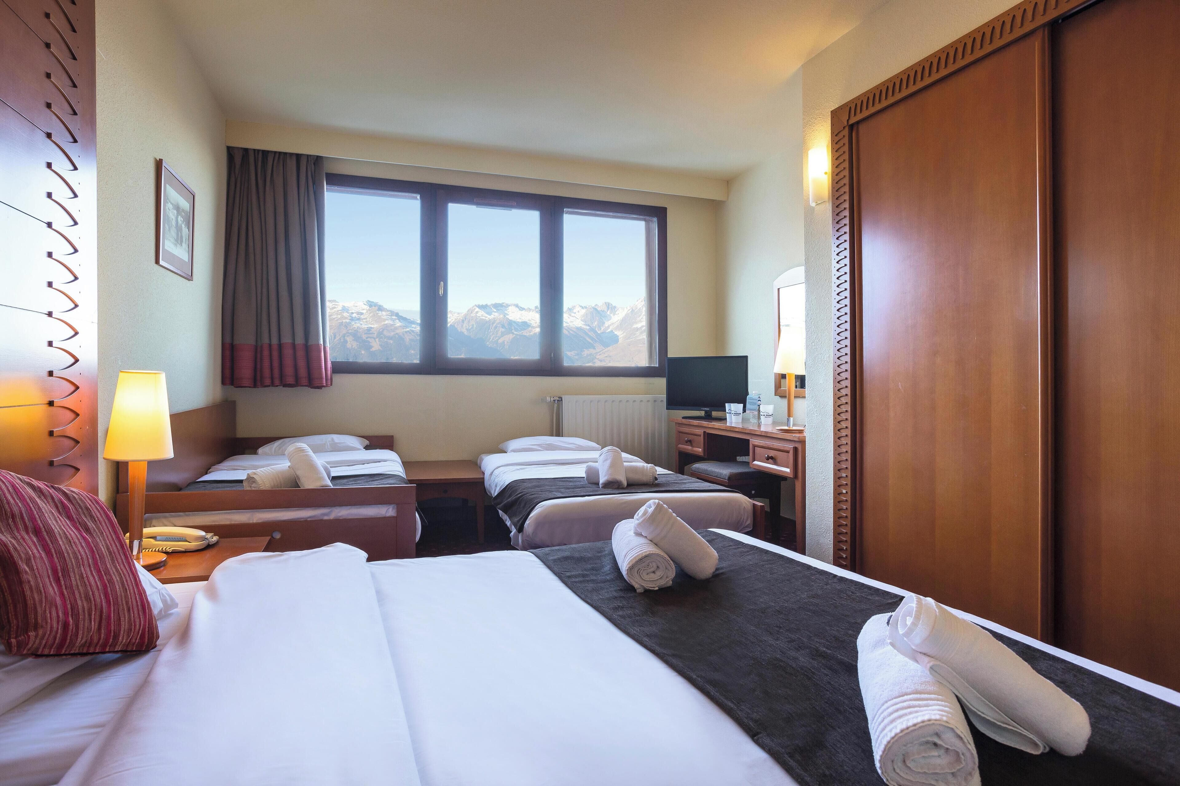 A hotel room with two beds, a desk, a lamp, a window with a mountain view, and a wardrobe.