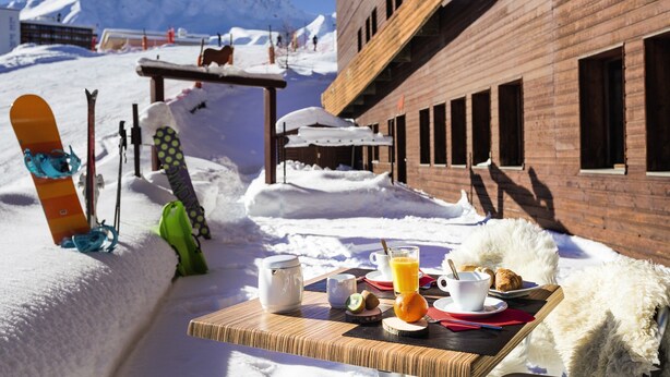 A breakfast setup with a snowboard and ski gear, a wooden table, and a building with a brick facade in a snowy outdoor setting.
