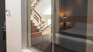 Habitación de hotel con una cama, una mesita de noche y una lámpara. Se ve una ventana con vistas a una escalera y una puerta.
