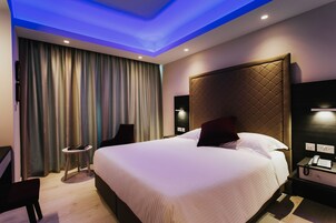 A modern hotel room with a large bed, a small side table, a chair, a TV, and a desk with a phone.
