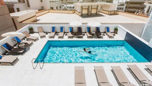 Piscina en la azotea con sillones de descanso y sombrillas, rodeada de edificios de la ciudad.