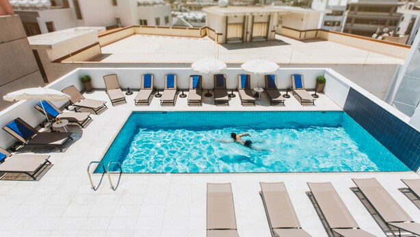 A rooftop swimming pool with lounge chairs and umbrellas, surrounded by city buildings.