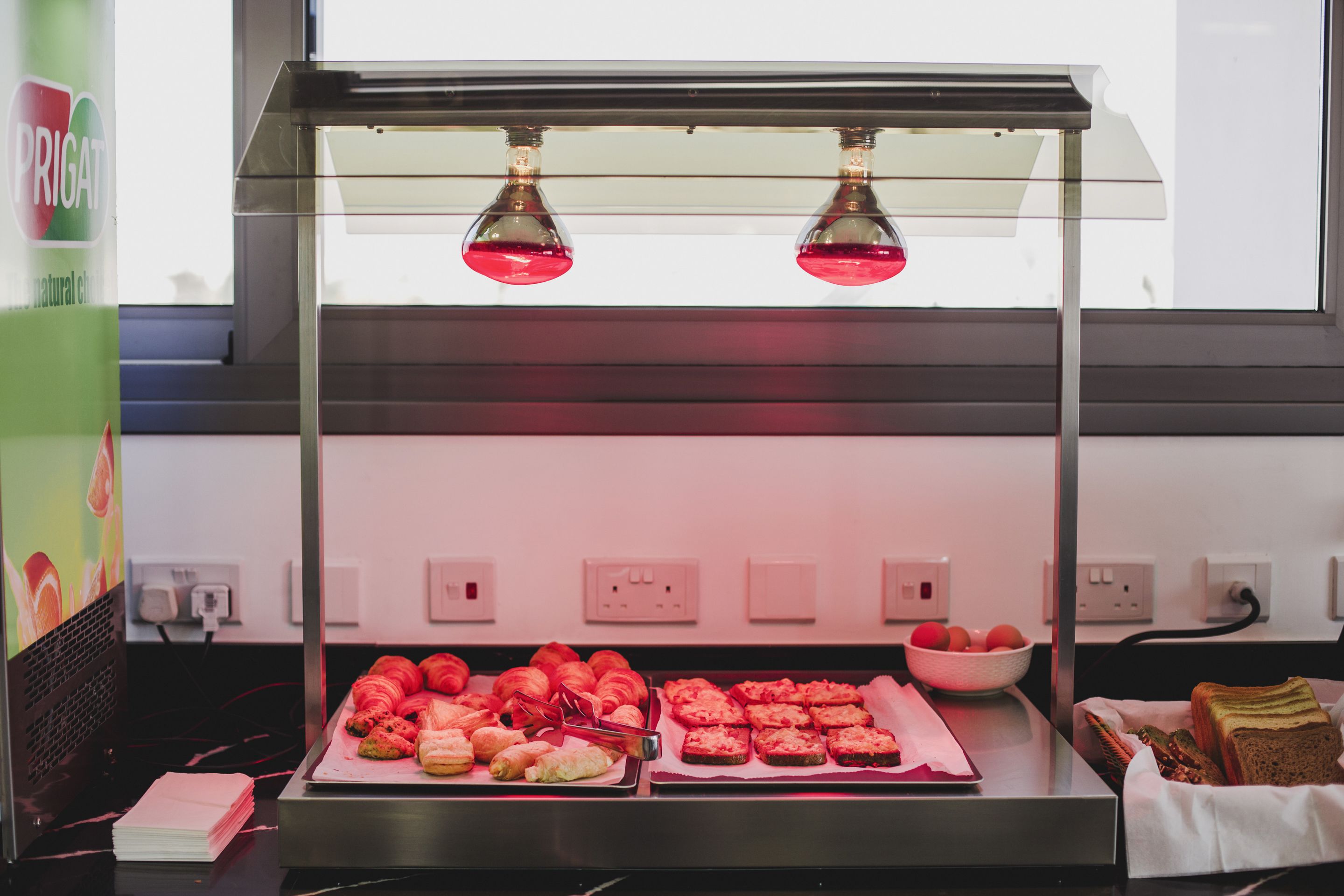 A food display with pastries and bread under a glass enclosure with two lights.