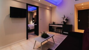 A modern hotel room with a flat-screen TV, a small table with a plant, a bed, a desk, and a bathroom visible through an open door.