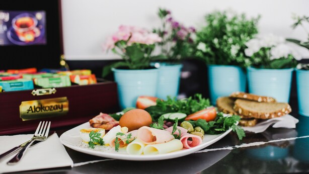 Un plato de comida con varios elementos, incluyendo carne, verduras y pan, un tenedor y una servilleta sobre una mesa. Hay una caja con el rótulo 'Alokozay' y una pantalla de televisión al fondo.