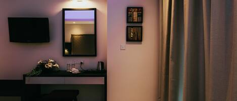 A hotel room with a bed, a desk with a mirror, a flat-screen TV, and a small shelf with decorative items.