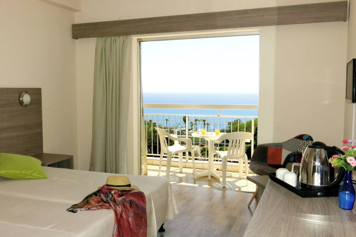 A hotel room with a bed, a small table with chairs, a balcony with a view of the ocean, and a coffee maker on the table.