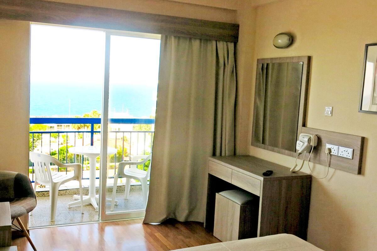A hotel room with a balcony, a desk, a chair, a bed, a TV, and a mirror.