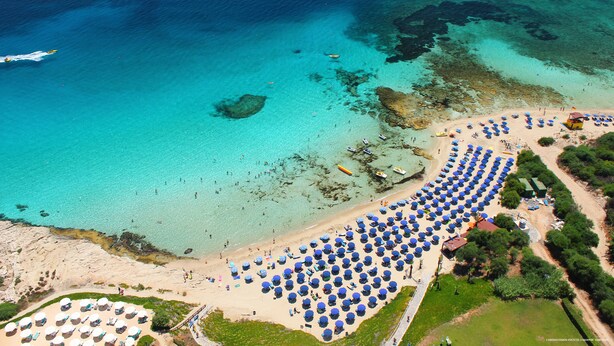 Vue aérienne d’une plage bordée de rangées de parasols et d’un rivage sablonneux.