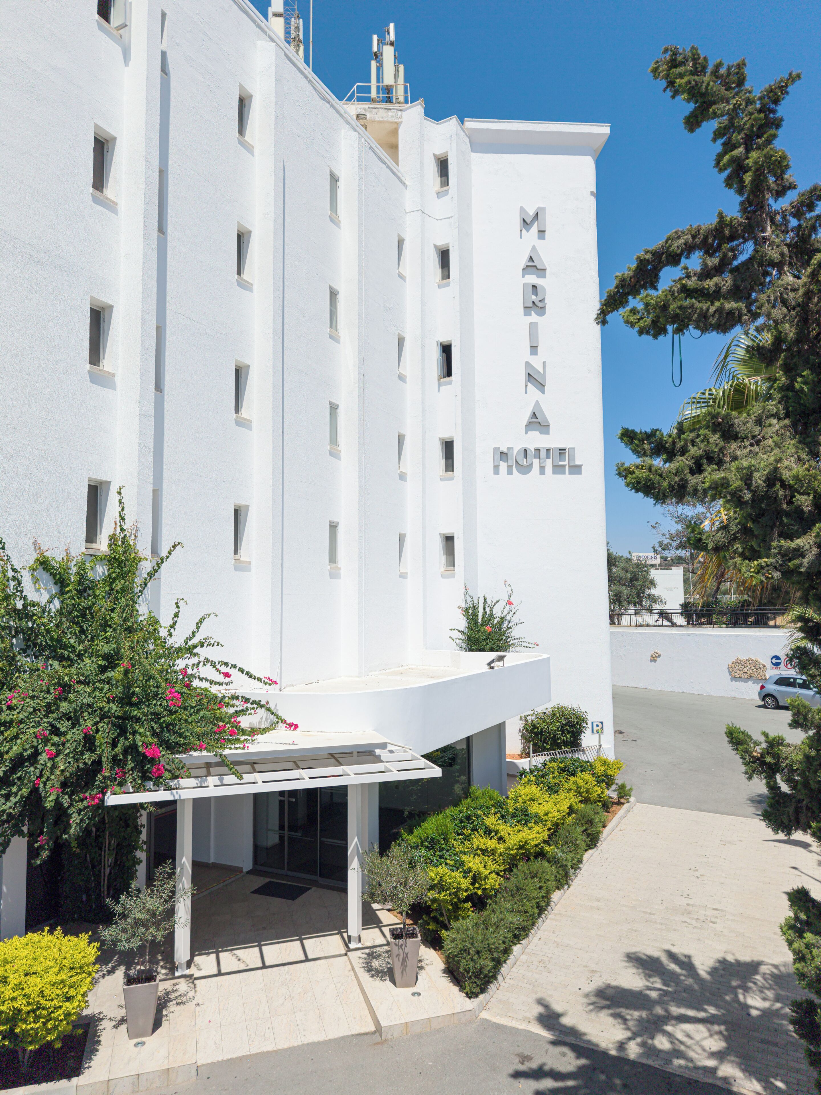 Un edificio blanco con el letrero 'MARINA HOTEL' en la fachada. Cuenta con un acceso cubierto y un pequeño jardín delantero.