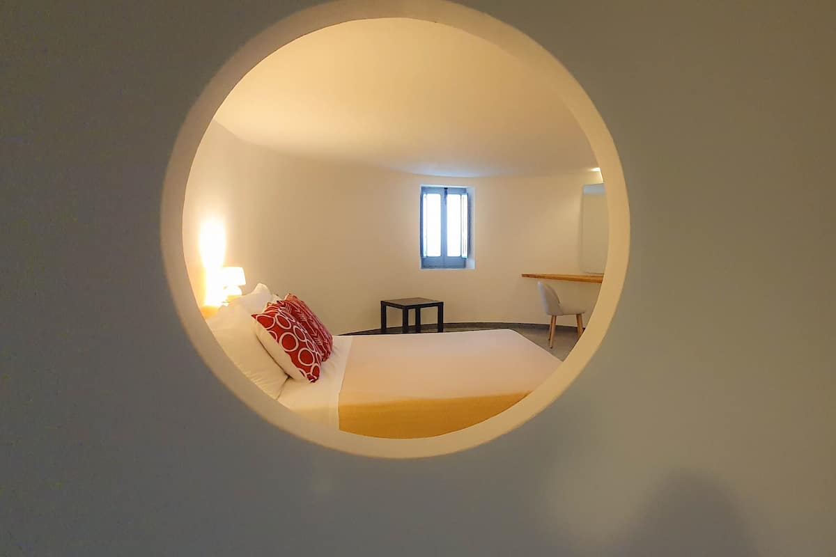 Een ronde spiegel die een kamer weerspiegelt met een bed, een kleine tafel, een stoel en een raam.