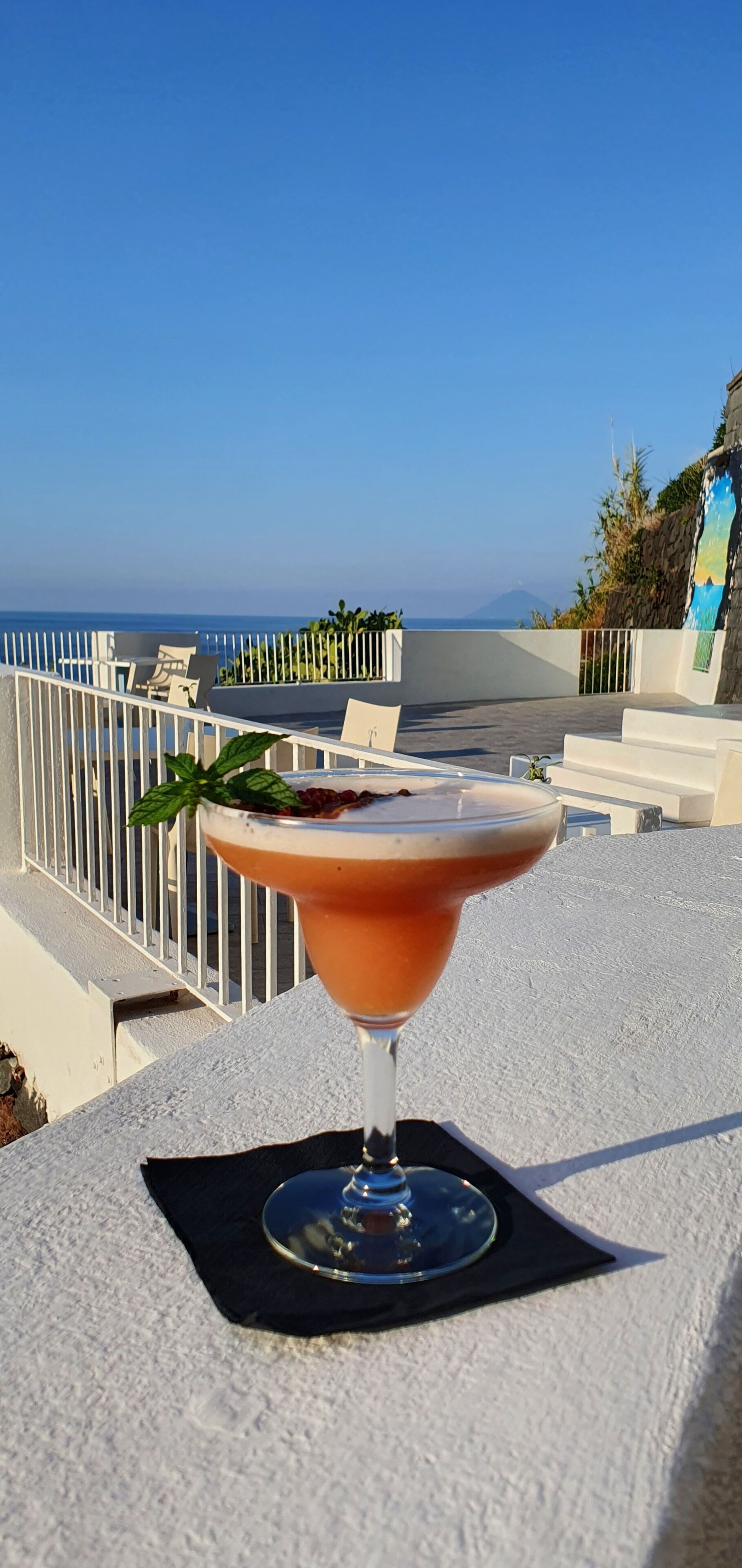Un cocktail sur un balcon avec vue sur la mer et une montagne à l’horizon.
