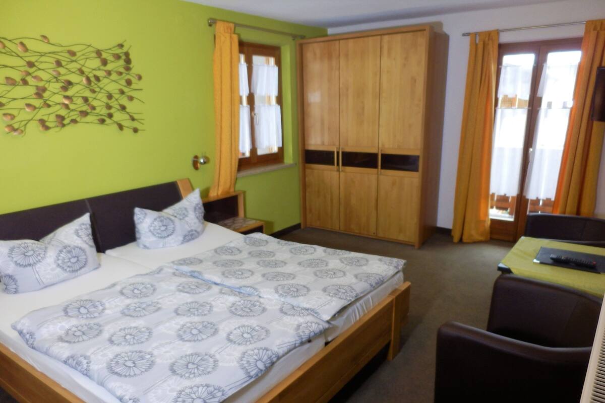 A bedroom with a bed, a wardrobe, a table with a chair, and a window with curtains.