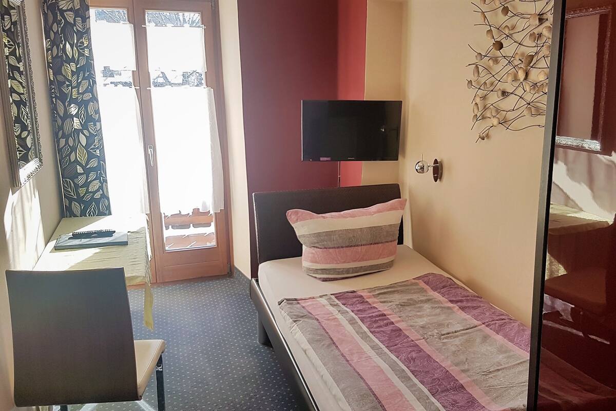 A bedroom with a bed, a desk, a chair, a TV, and a door.