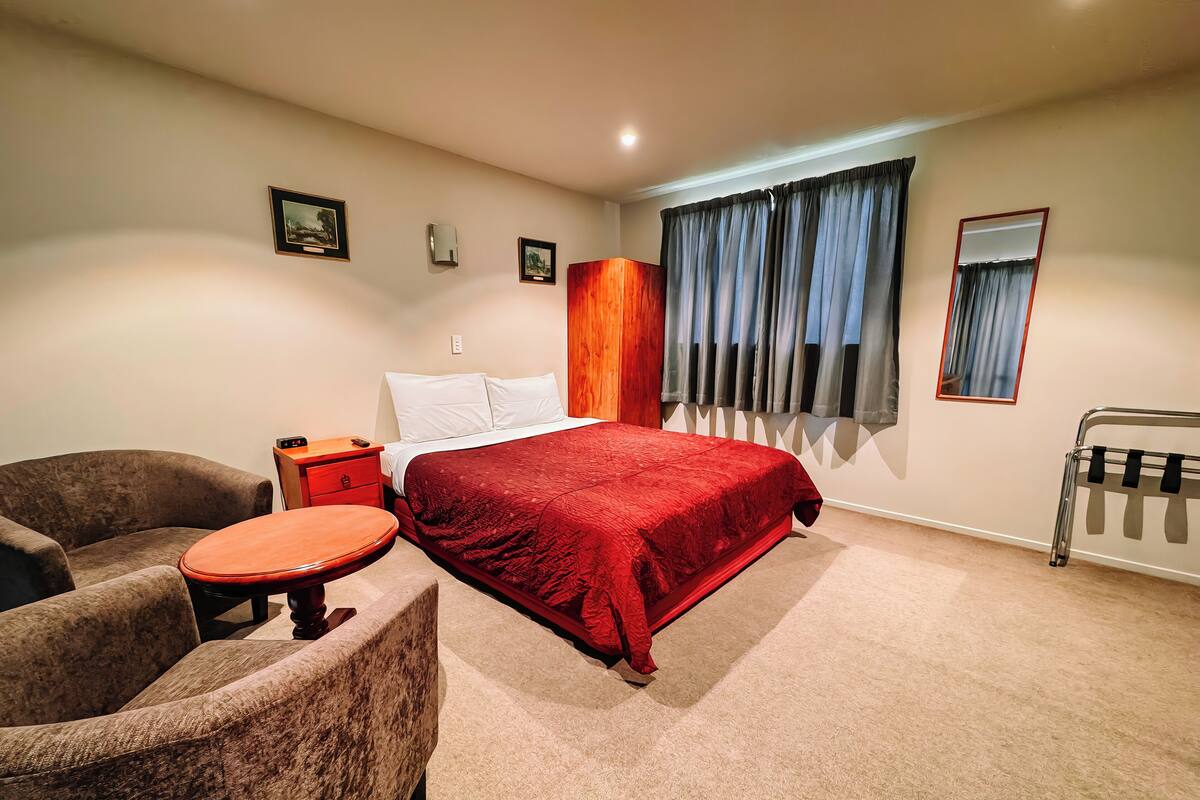 A hotel room with a large bed, a wooden nightstand, a chair, a small table, and a radiator.
