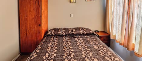 Une chambre avec une armoire en bois, un lit recouvert d’une couvre-lit à motifs de feuilles, une table de chevet, une petite table d’appoint, une lampe fixée au mur et un tableau encadré accroché au mur.