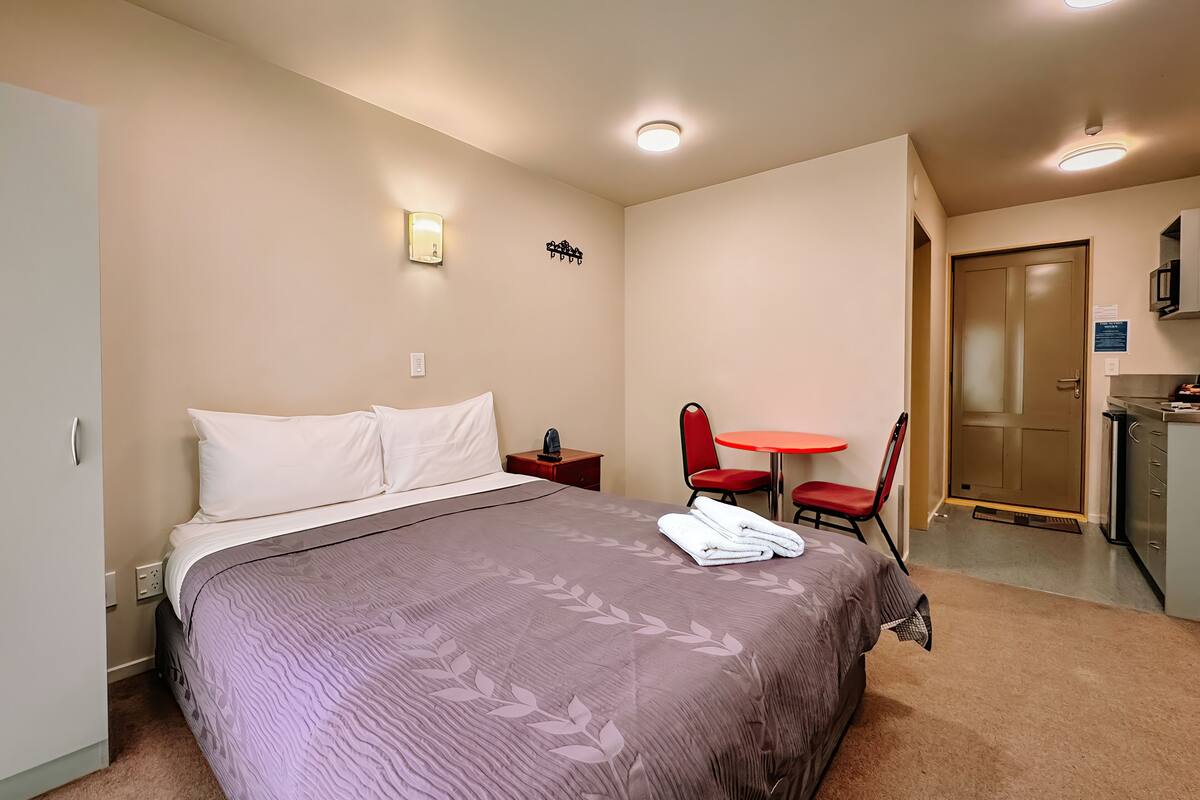 A neatly arranged hotel room with a bed, a small table with chairs, a desk, and a door.