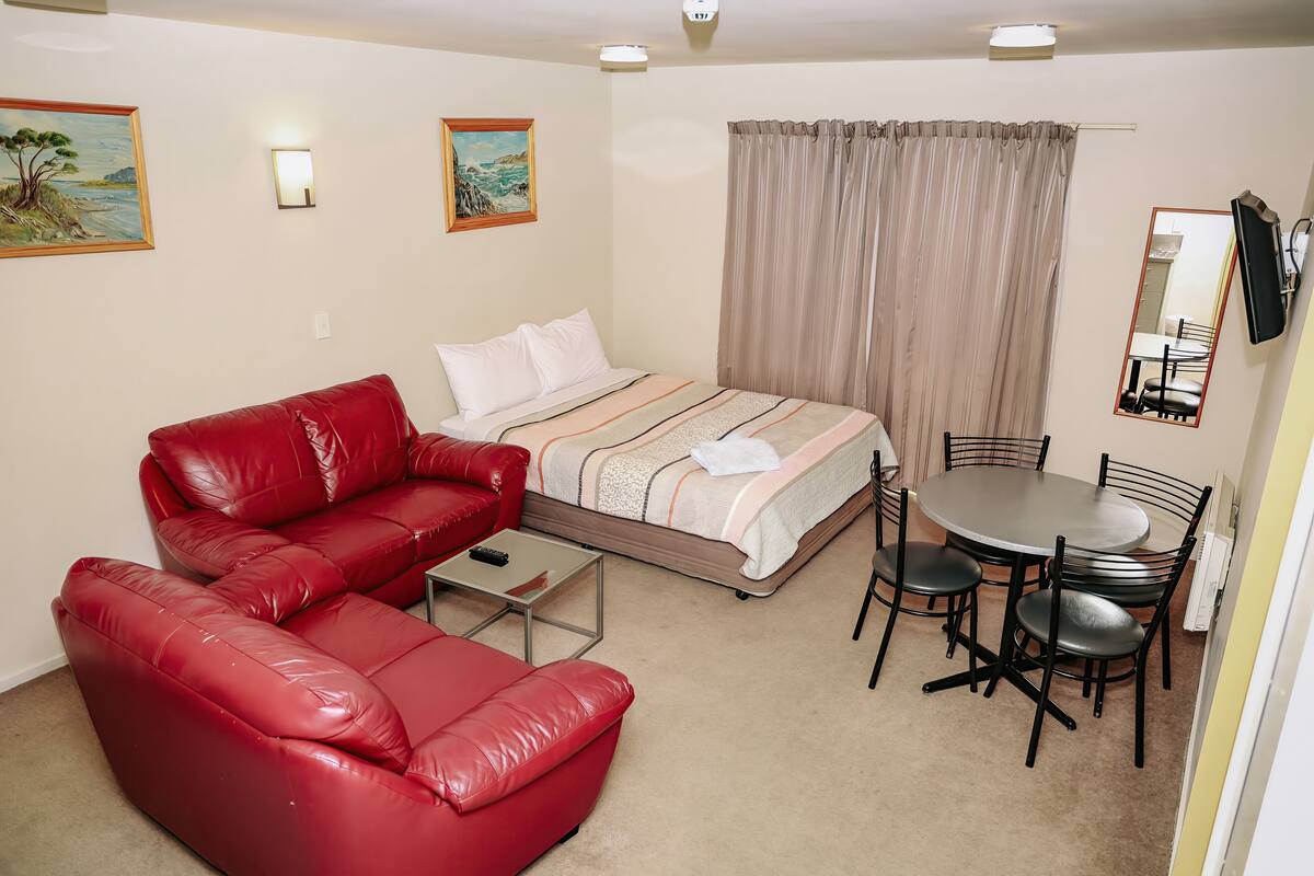 A room with a bed, a red leather sofa, a small table, a round dining table with chairs, and two wall-mounted mirrors.