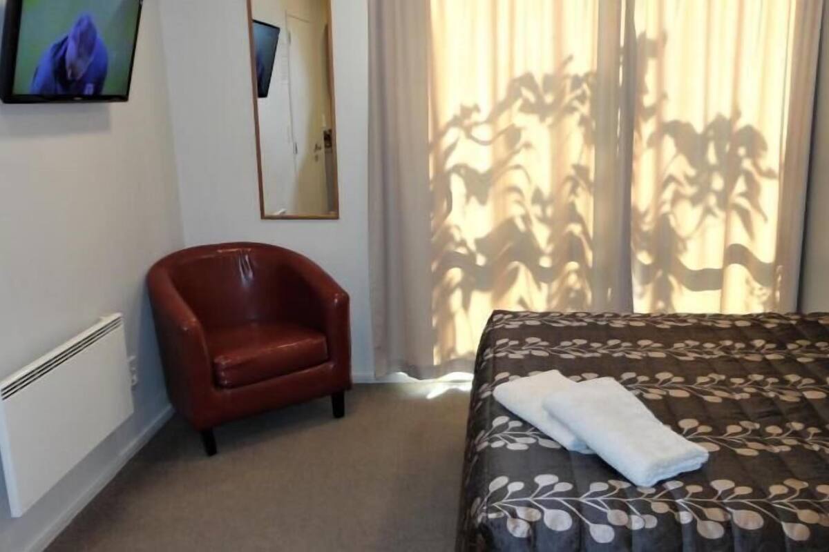 A hotel room with a bed, a red armchair, a large window with curtains, and a wall-mounted TV.
