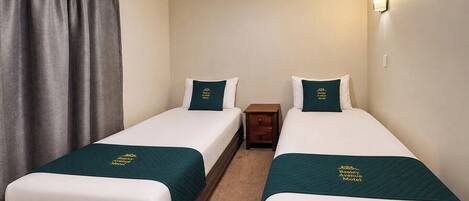 Habitación de hotel con dos camas individuales, cada una con cubrecama y almohada verde a juego, una mesita de noche de madera y una lámpara fijada a la pared.