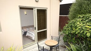 Un pequeño patio con una mesa y sillas, una puerta corrediza de vidrio que da a un dormitorio y un arbusto verde al fondo.