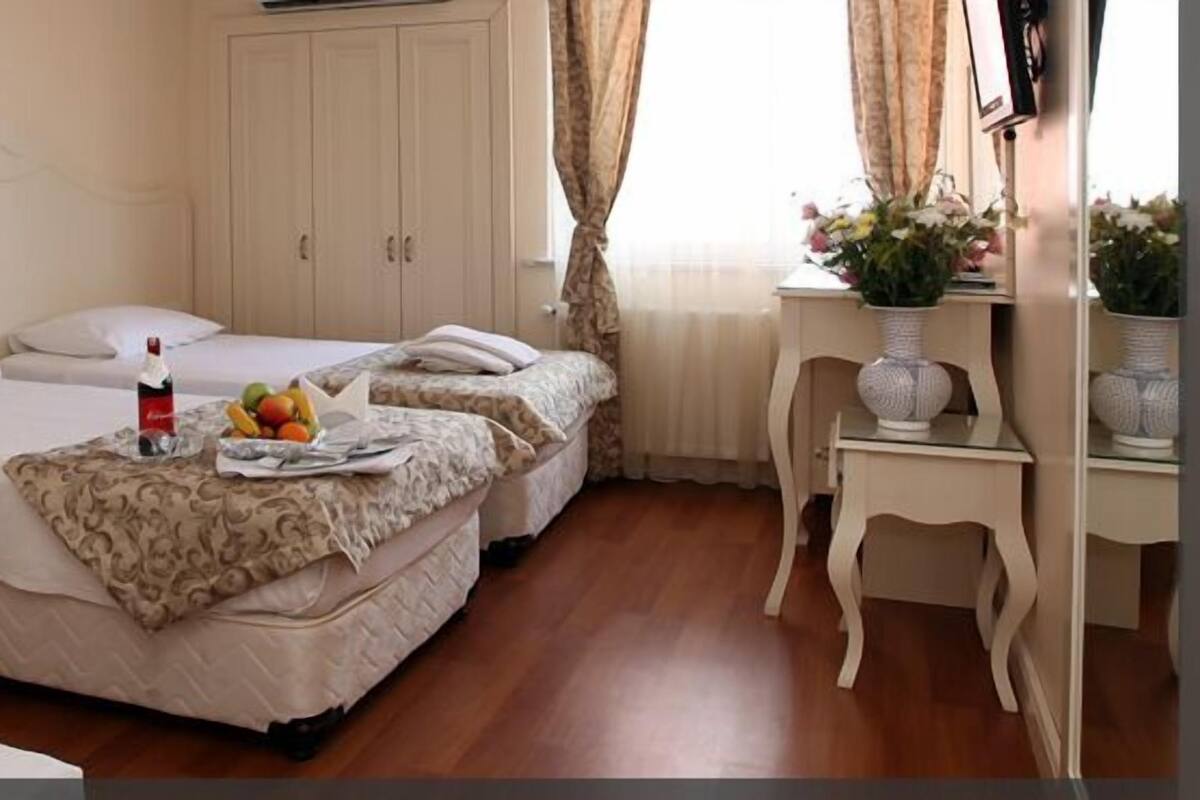 A hotel room with two beds, a bedside table with a lamp, a small table with a vase of flowers, and a fruit basket on one of the beds.