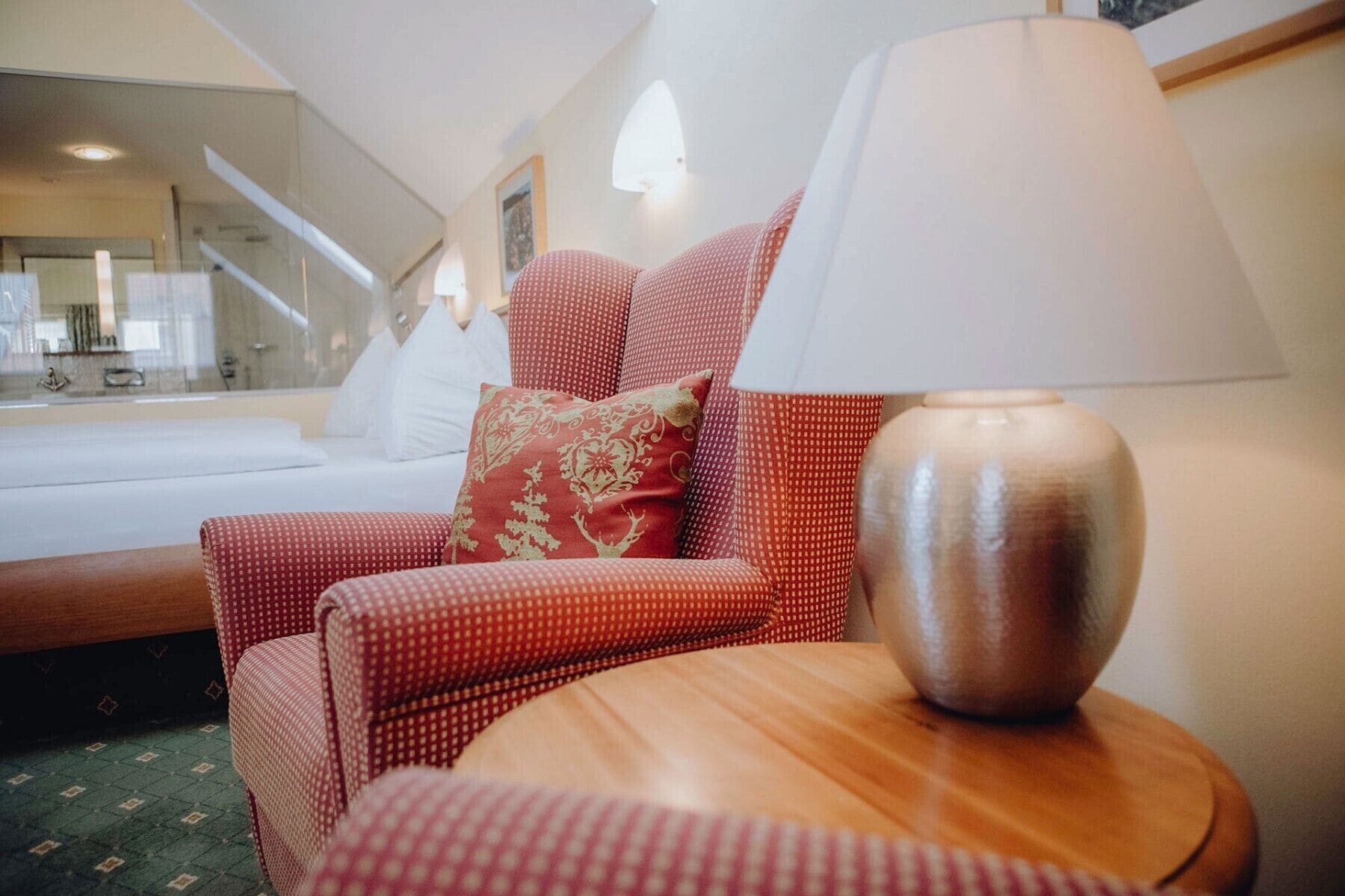 A hotel room with a bed, a red upholstered chair with a patterned cushion, a wooden side table with a lamp, and a large mirror reflecting the room.