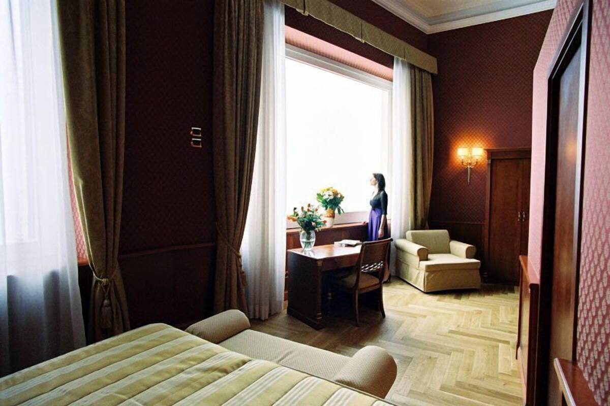 Une chambre d’hôtel avec un lit, un bureau et une chaise, une petite table avec un vase, et une fenêtre avec des rideaux.