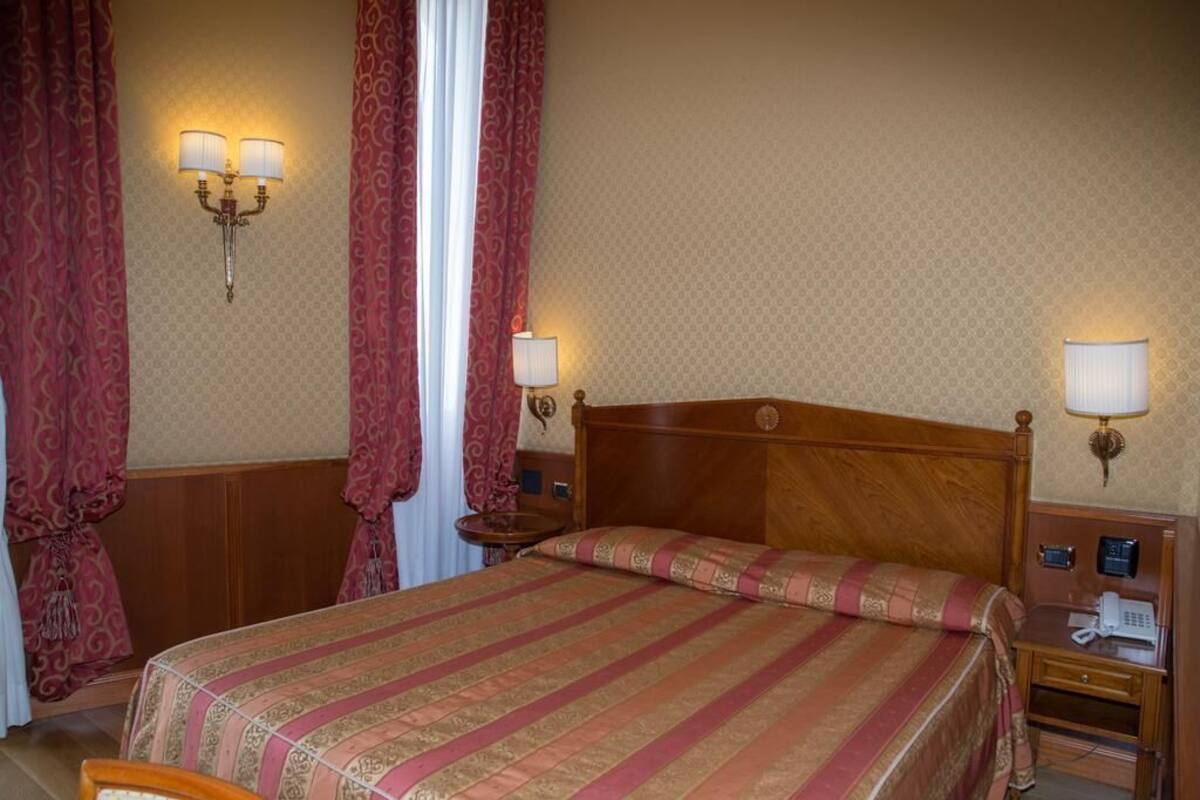 Une chambre d’hôtel avec un lit en bois, deux lampes de chevet, une table de chevet avec un téléphone et des rideaux à motifs.