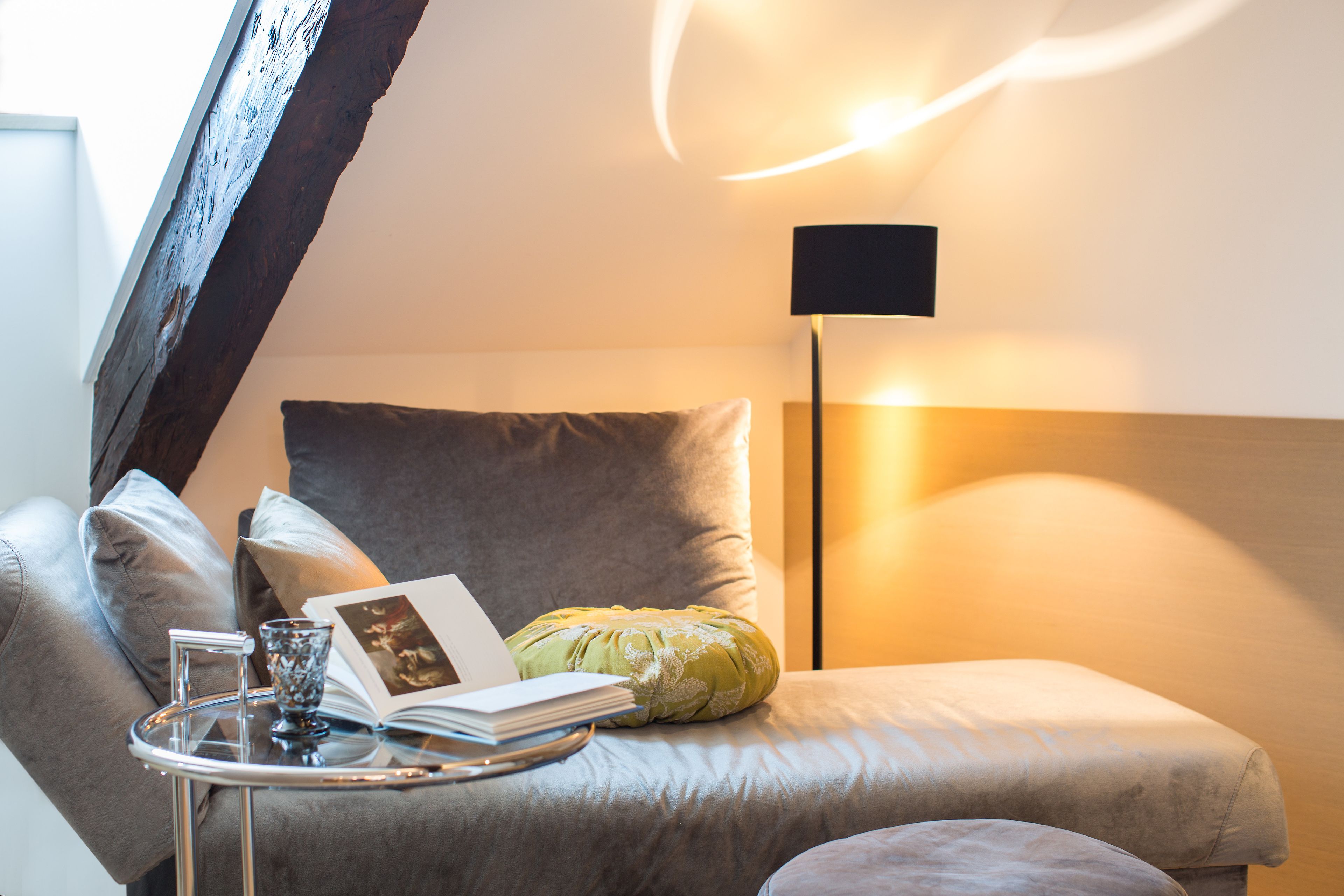 A cozy reading nook with a cushioned chair, a small table with a book and a glass, and a floor lamp.