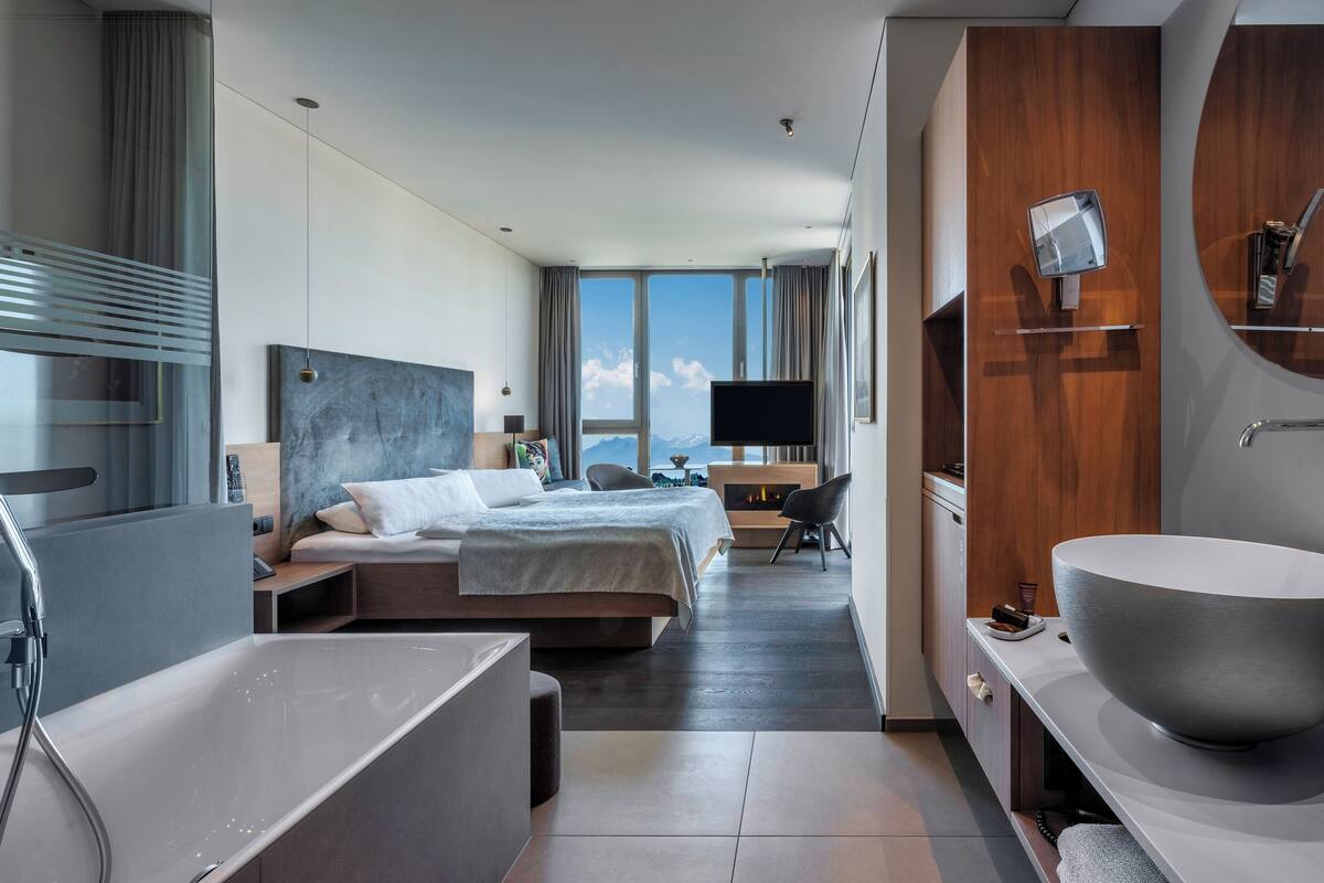 A modern hotel room with a large bed, a TV, a fireplace, a large mirror, and a large sink.