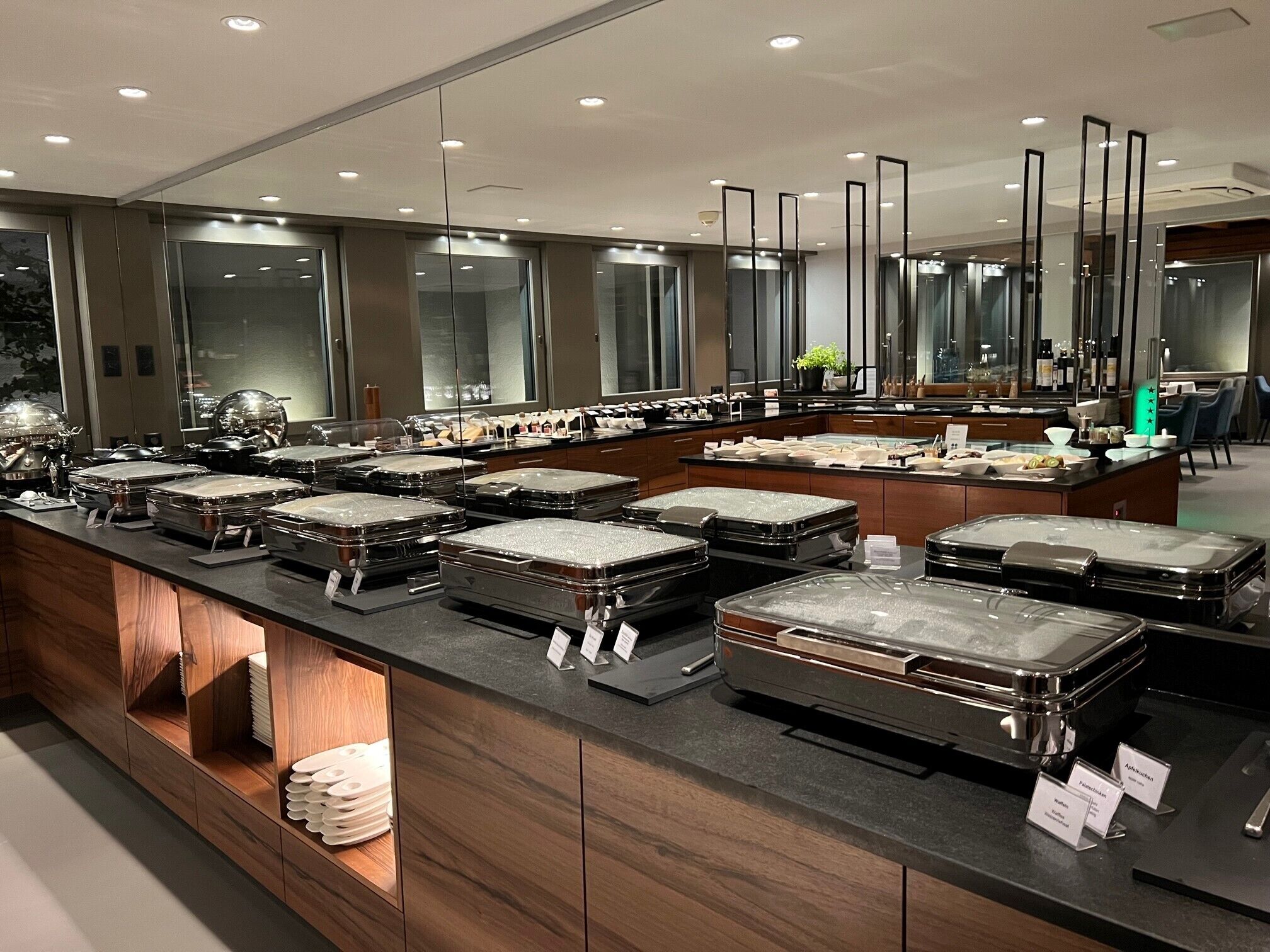 A buffet area with multiple serving stations, each with a covered food container. There are wooden cabinets below the buffet, and a large mirror above the counter.