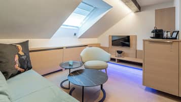 Ein modernes Schlafzimmer im Loft-Stil mit Dachfenster, einer Couch, einem Sessel, einem kleinen runden Tisch und einem an der Wand montierten Fernseher.