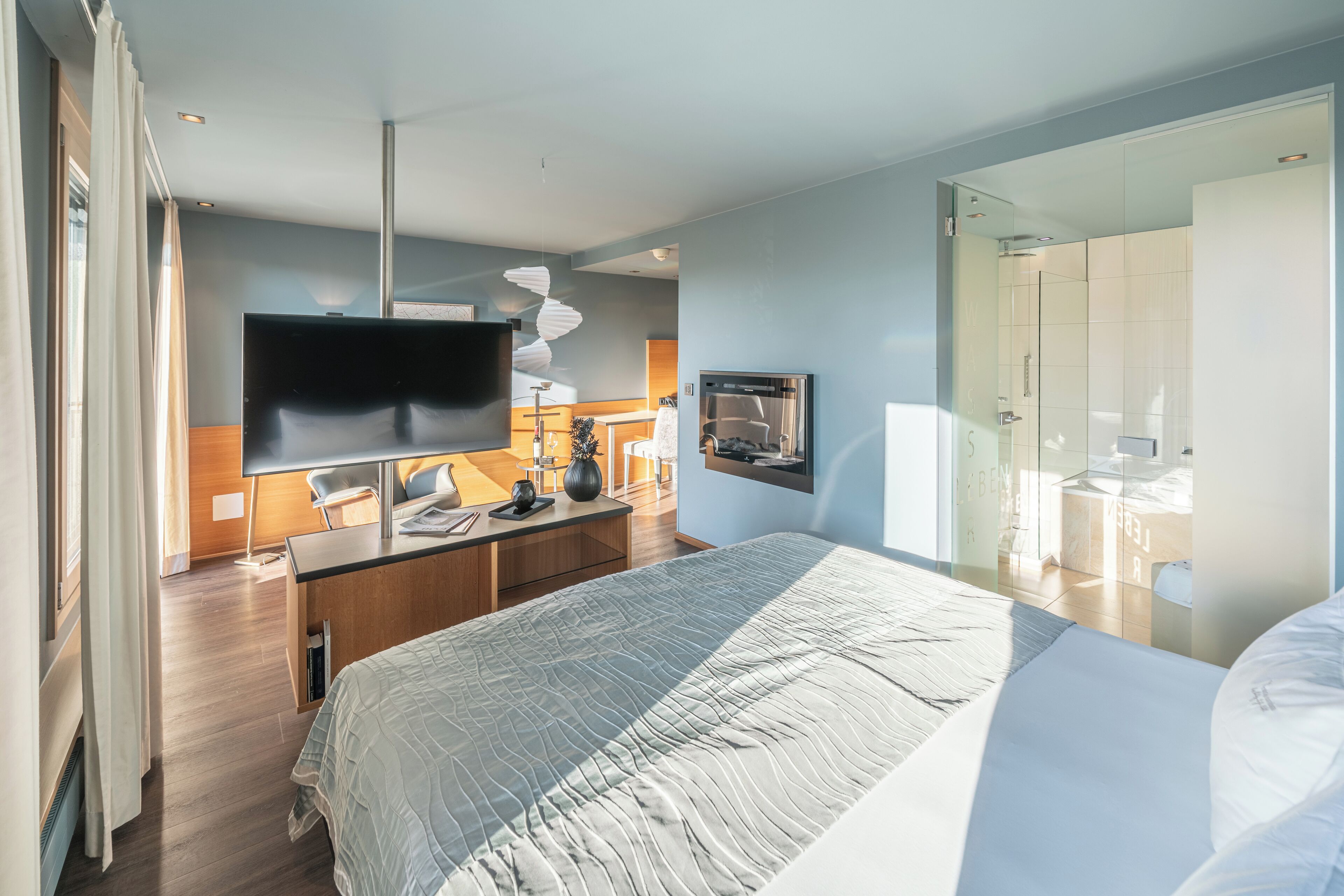 A modern hotel room with a large bed, a desk with a flat-screen TV, a refrigerator, and a glass-enclosed bathroom.