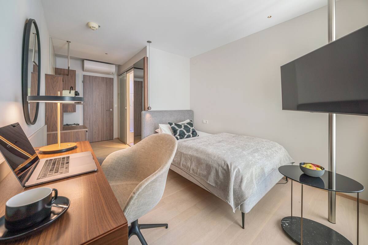 A modern hotel room with a bed, a desk with a laptop, a chair, a TV, and a small table with a bowl of fruit.