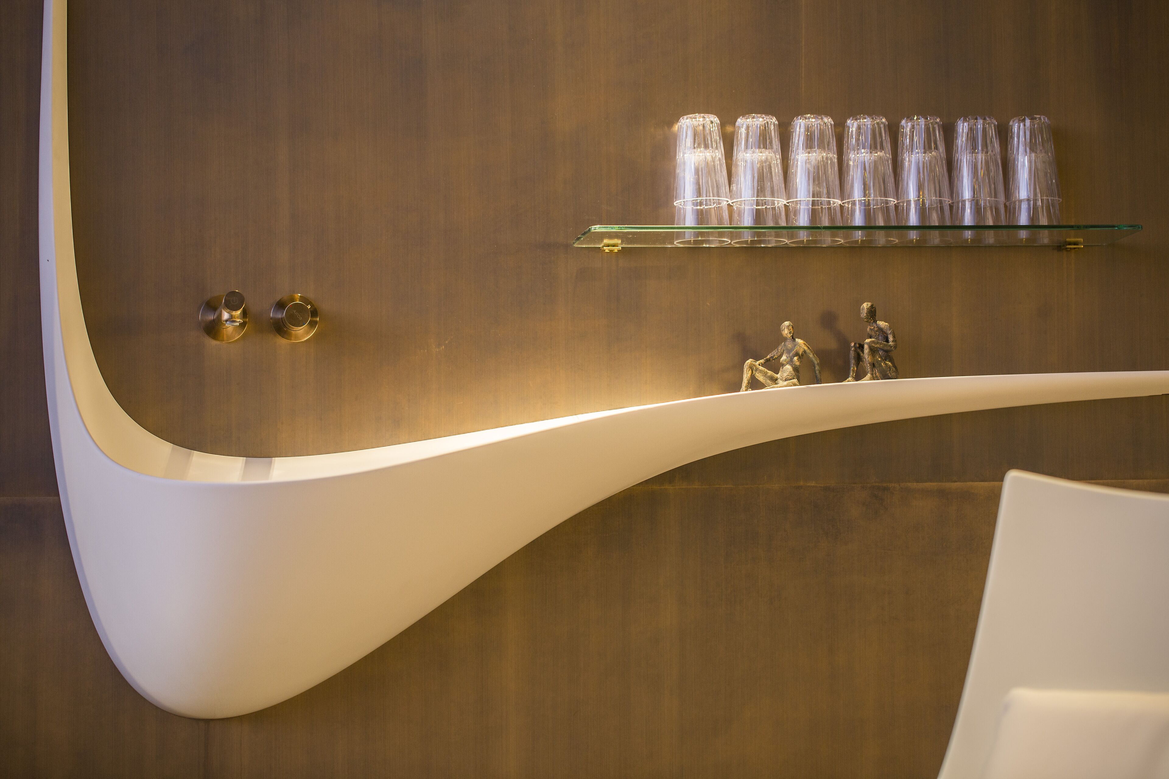 A modern bathroom with a curved white sink, a wooden wall, and a glass shelf holding clear glasses and a small sculpture.
