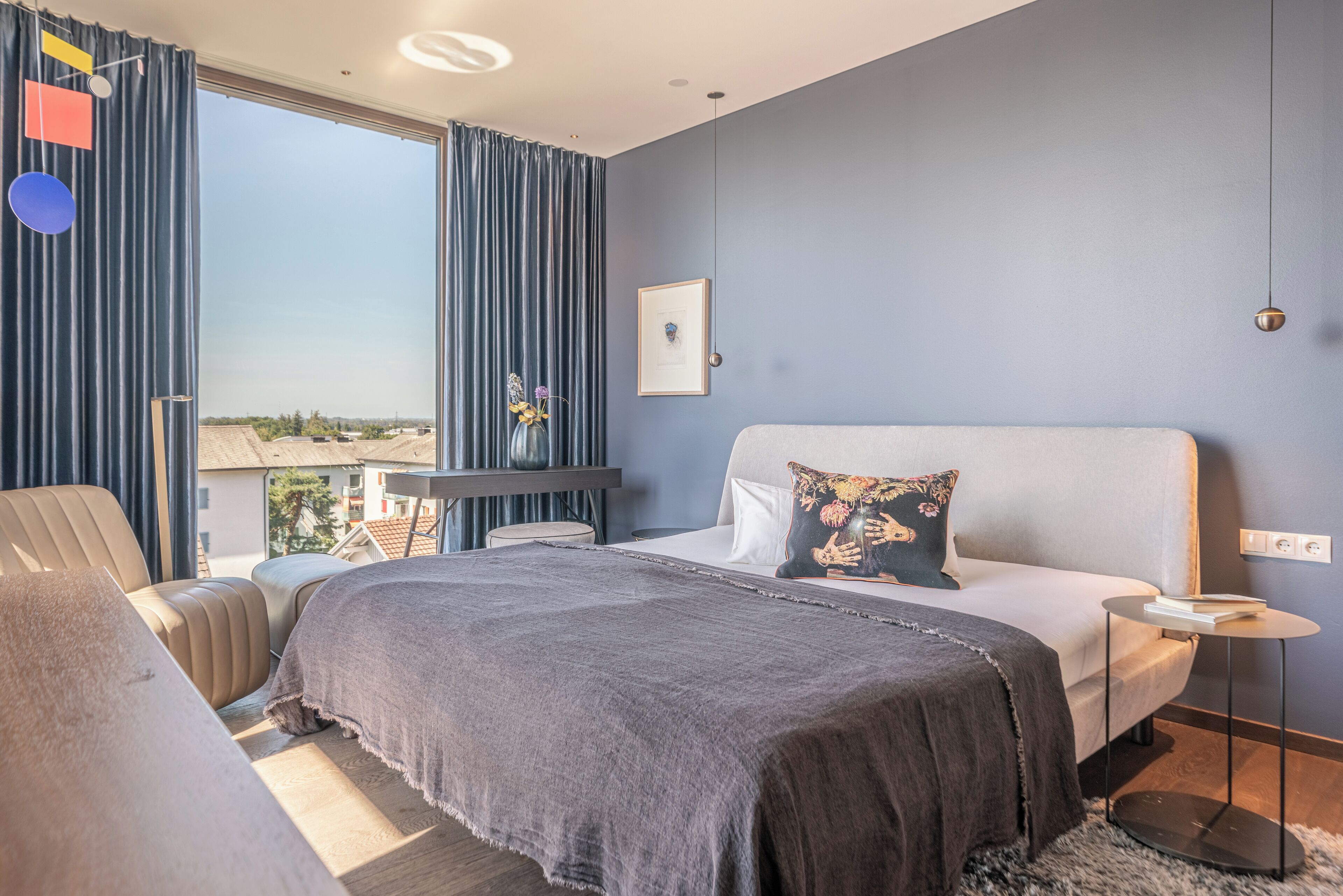 Ein modernes Schlafzimmer mit einem großen Bett, einem Schreibtisch und einem Stuhl. Ein Fenster mit Vorhängen, ein gerahmtes Bild an der Wand und ein Blick nach draußen.