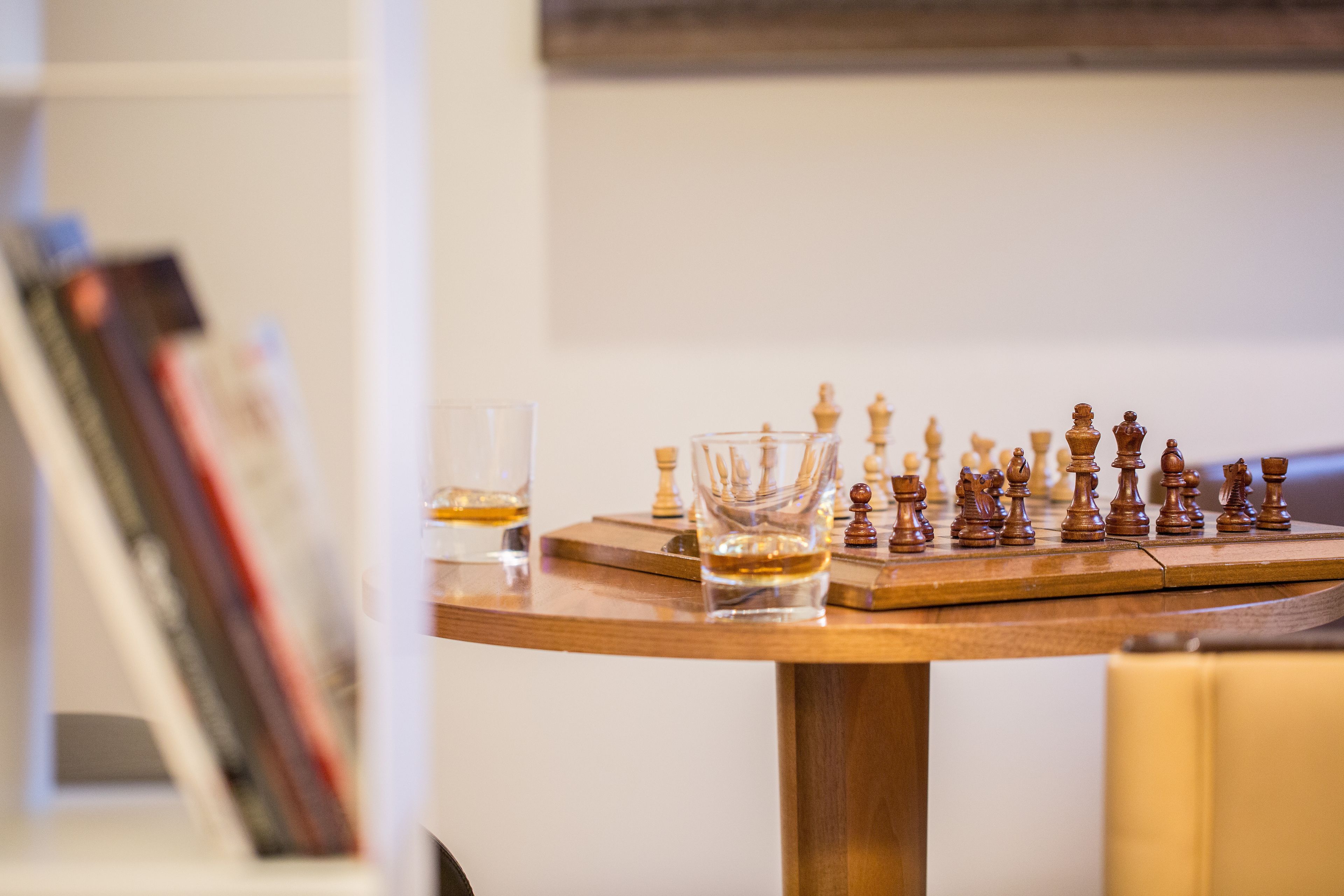 A chessboard on a wooden table with glasses and books in the background.