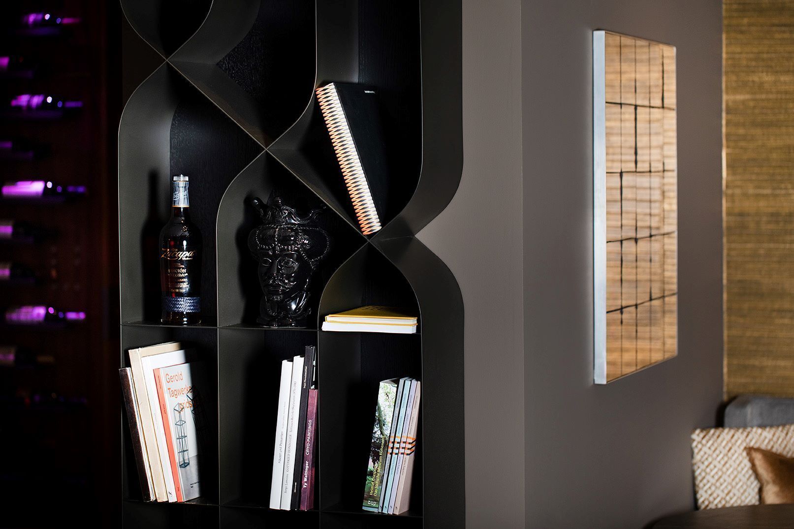 Un interior moderno con una estantería negra que contiene libros, una botella y un objeto decorativo. Se observa una obra de arte enmarcada en la pared.
