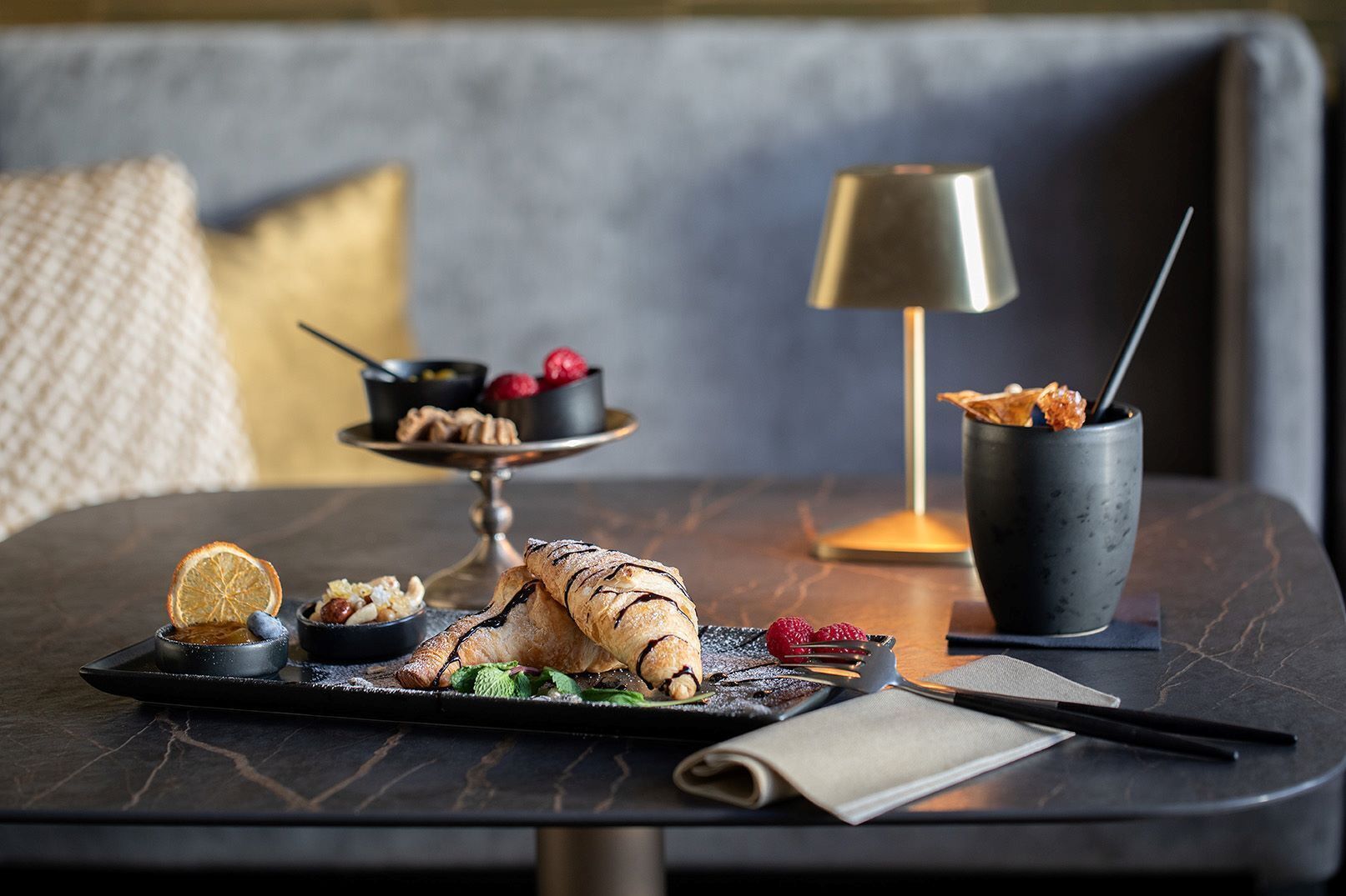A dining table with a croissant, a bowl of raspberries, a small dish with nuts, and a candle holder.