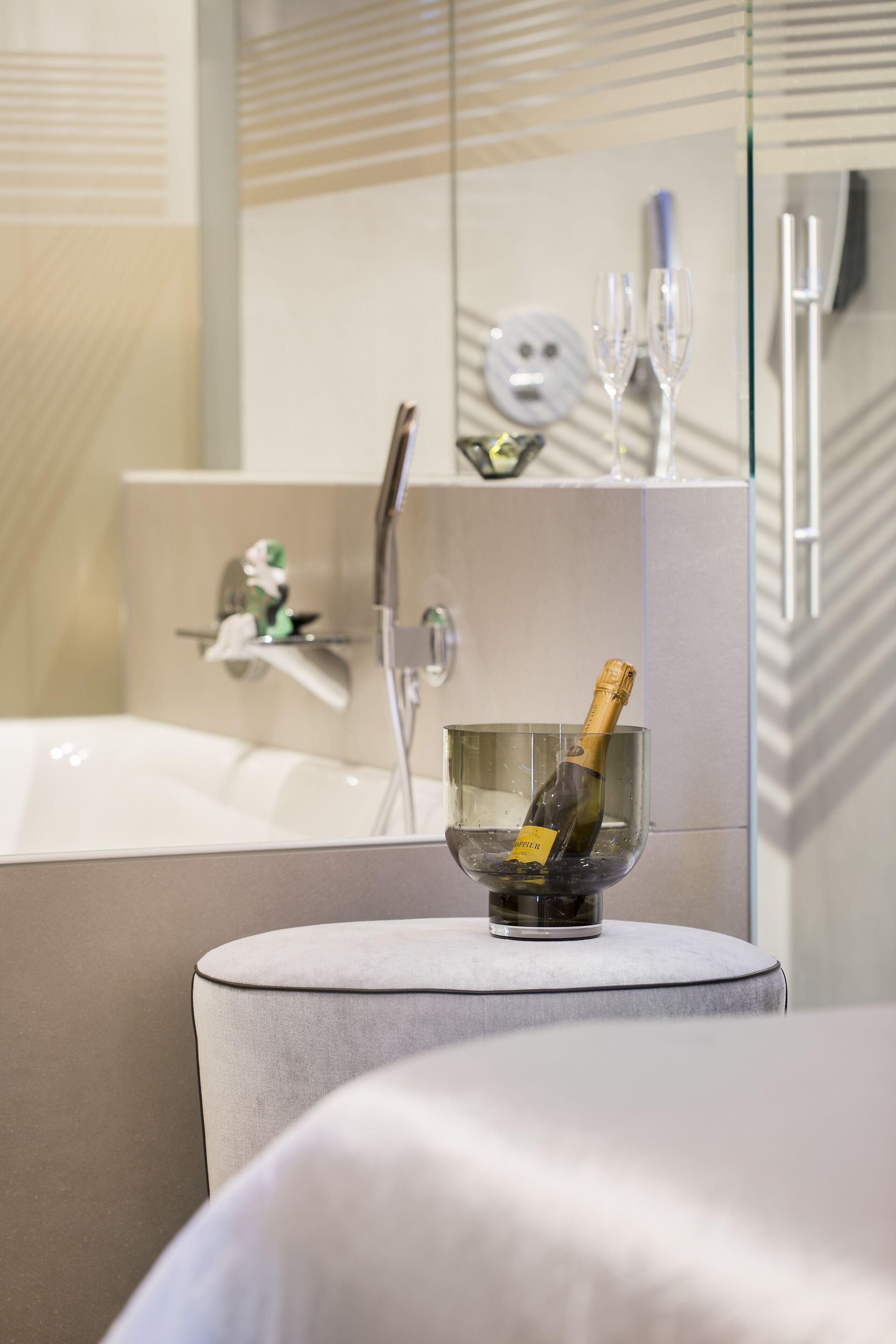 A modern bathroom with a bathtub, a glass bowl with a bottle of champagne, and a mirror reflecting a toothbrush and a wine glass.