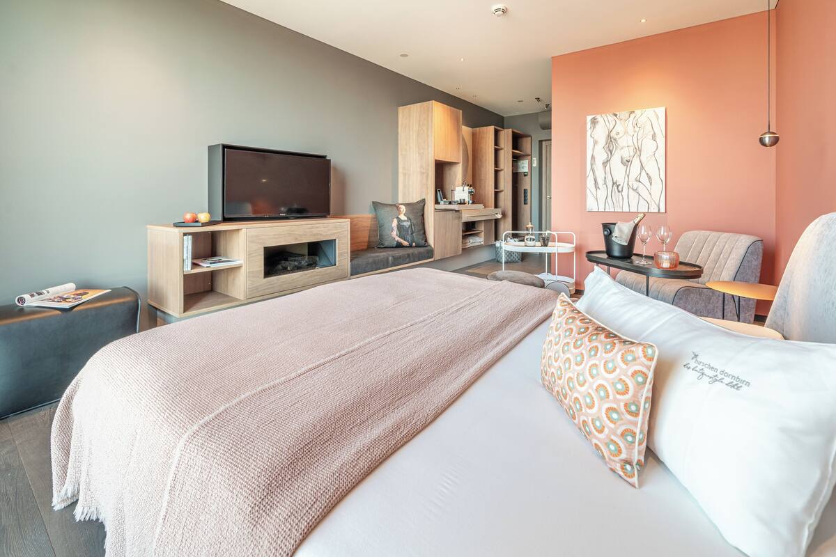 A modern hotel room with a large bed, a flat-screen TV, a seating area with a table, and a kitchenette.