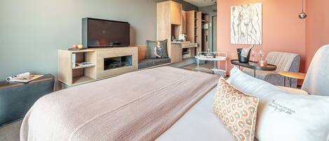 A modern hotel room with a large bed, a flat-screen TV, a seating area with a table, and a kitchenette.