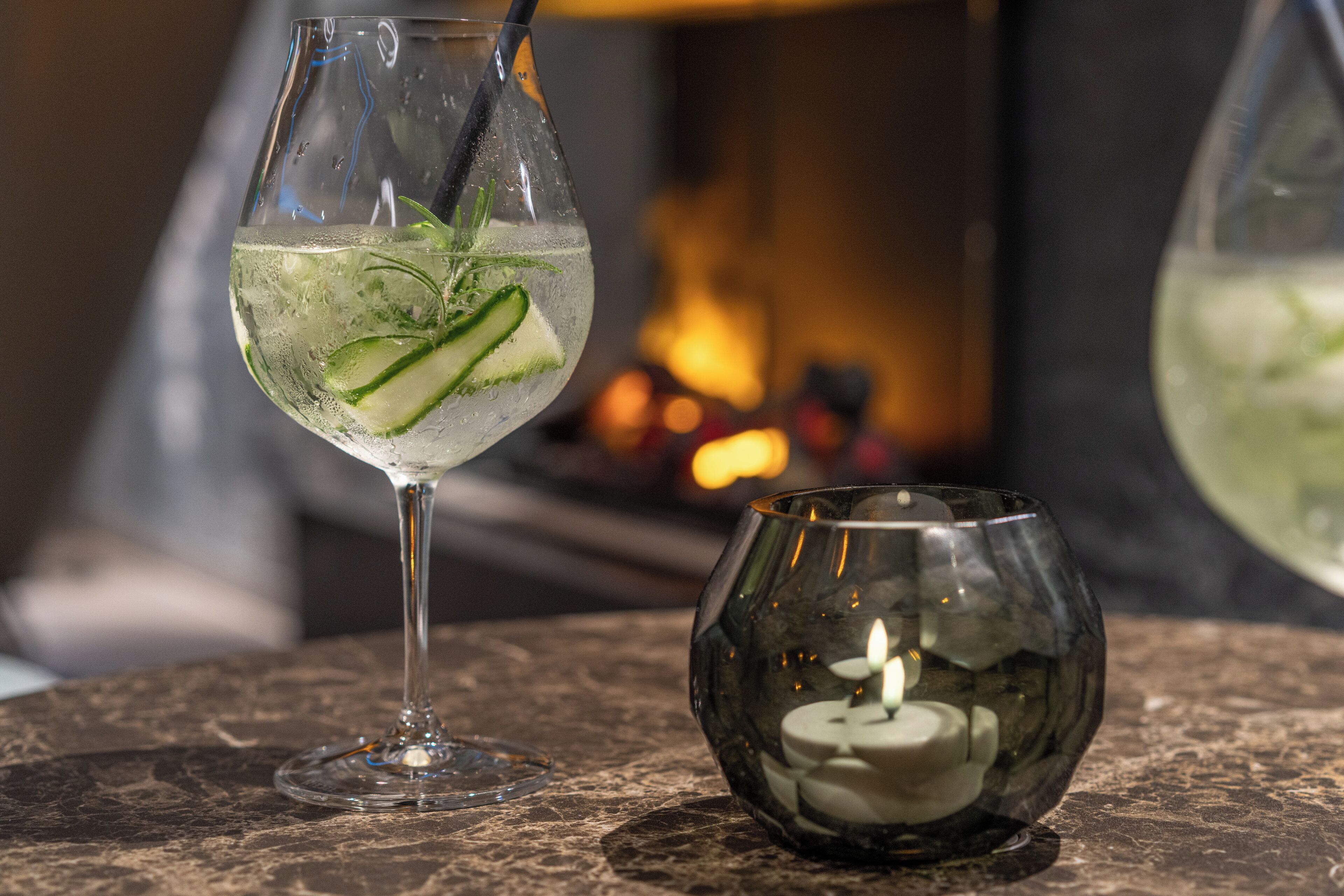 Two glasses of a clear beverage with slices of cucumber, a candle in a glass container, and a blurred background with a warm light source.