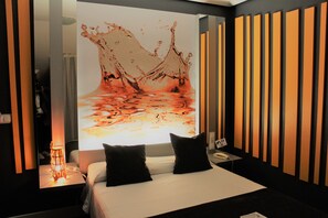 Habitación de hotel moderna con una gran obra de arte digital en la pared, una cama con almohadones negros, una mesita de noche con lámpara y una pared con listones de madera verticales.