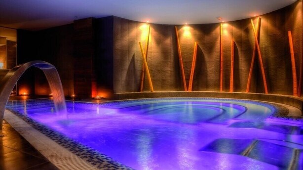 Un jacuzzi con diseño curvo, iluminado con luces cálidas y frías, y rodeado por un camino de baldosas.