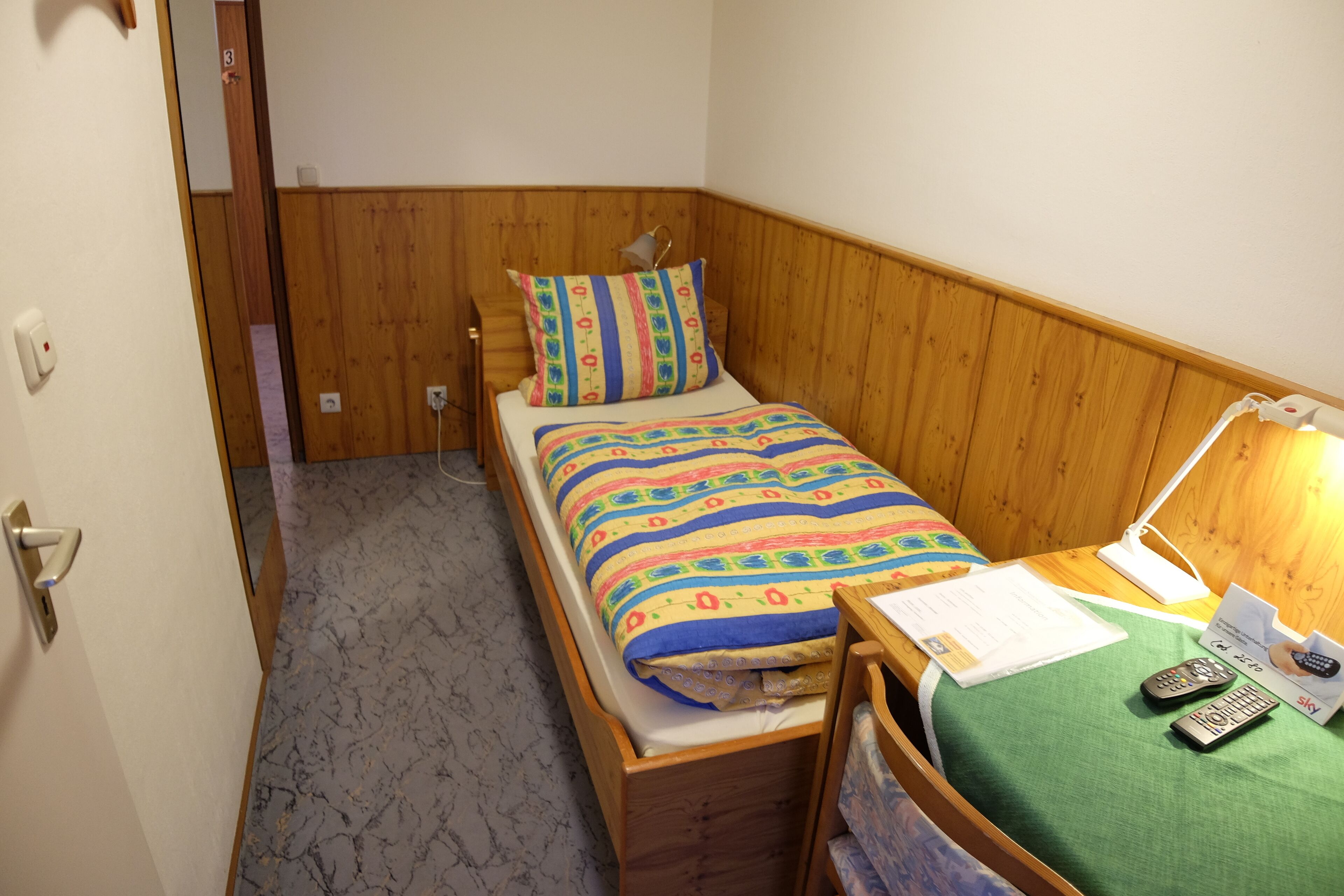 A small, single-bed room with wooden paneling, a green bedspread, a bedside lamp, and two remote controls on the bed.
