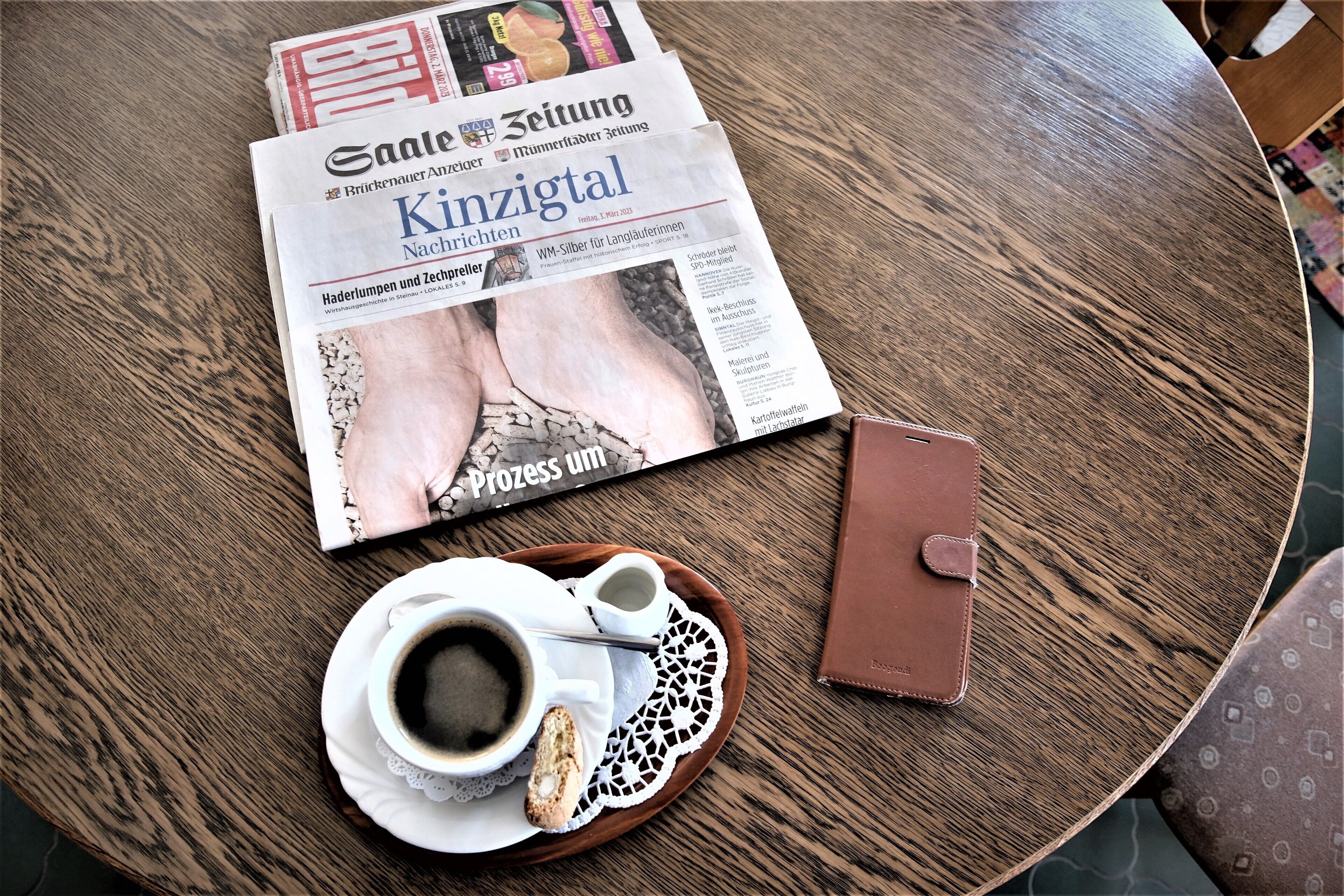 Una mesa con un periódico, una taza de café, un pastel y una billetera.