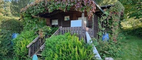 Una cabaña de madera con un porche, rodeada de abundante vegetación y una pequeña escalera que lleva a la entrada.